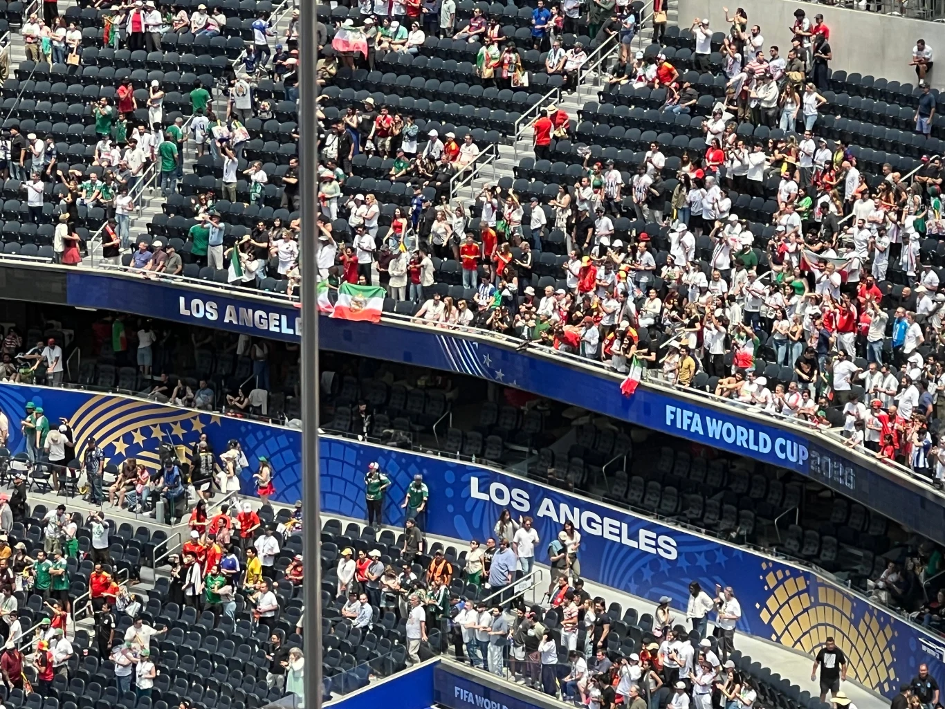 Tłum kibiców w białych i czerwonych koszulkach wypełnia trybuny stadionu oznaczonego jako Los Angeles podczas wydarzenia FIFA World Cup, wiele osób stoi i trzyma narodowe flagi.