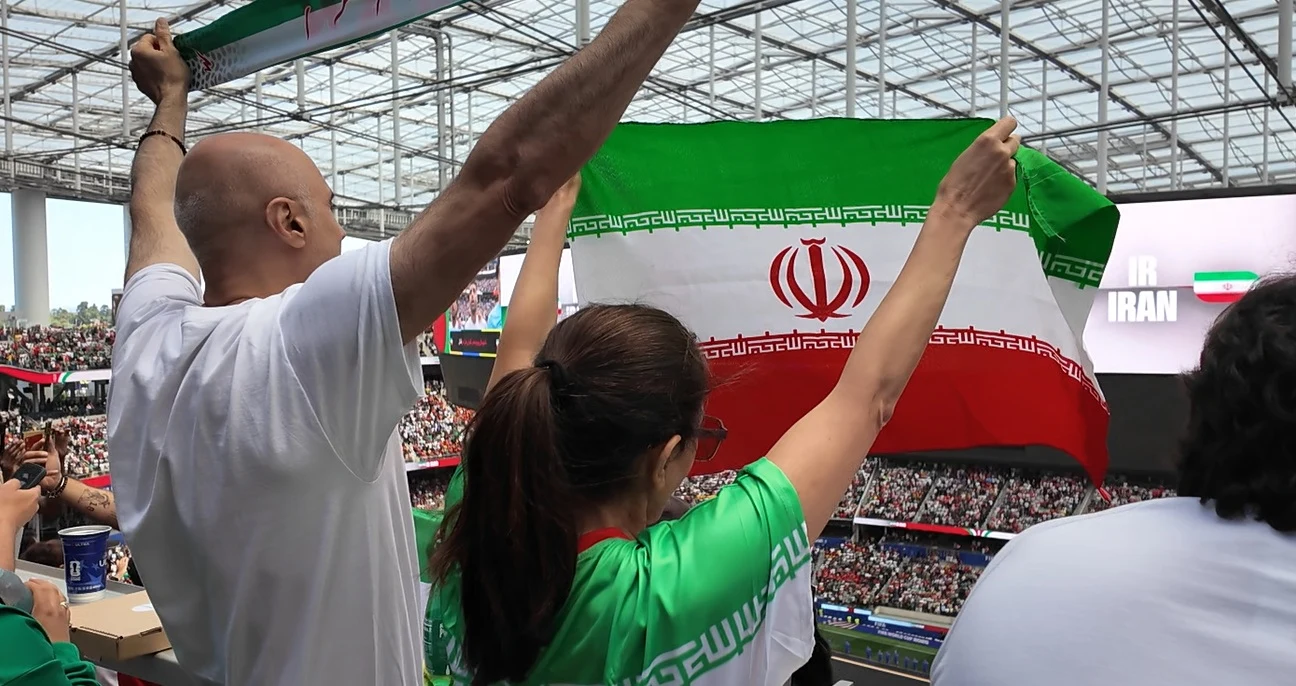 Kibice z flagą Iranu wznoszą ręce podczas meczu na stadionie, wypełnionym tłumem widzów.