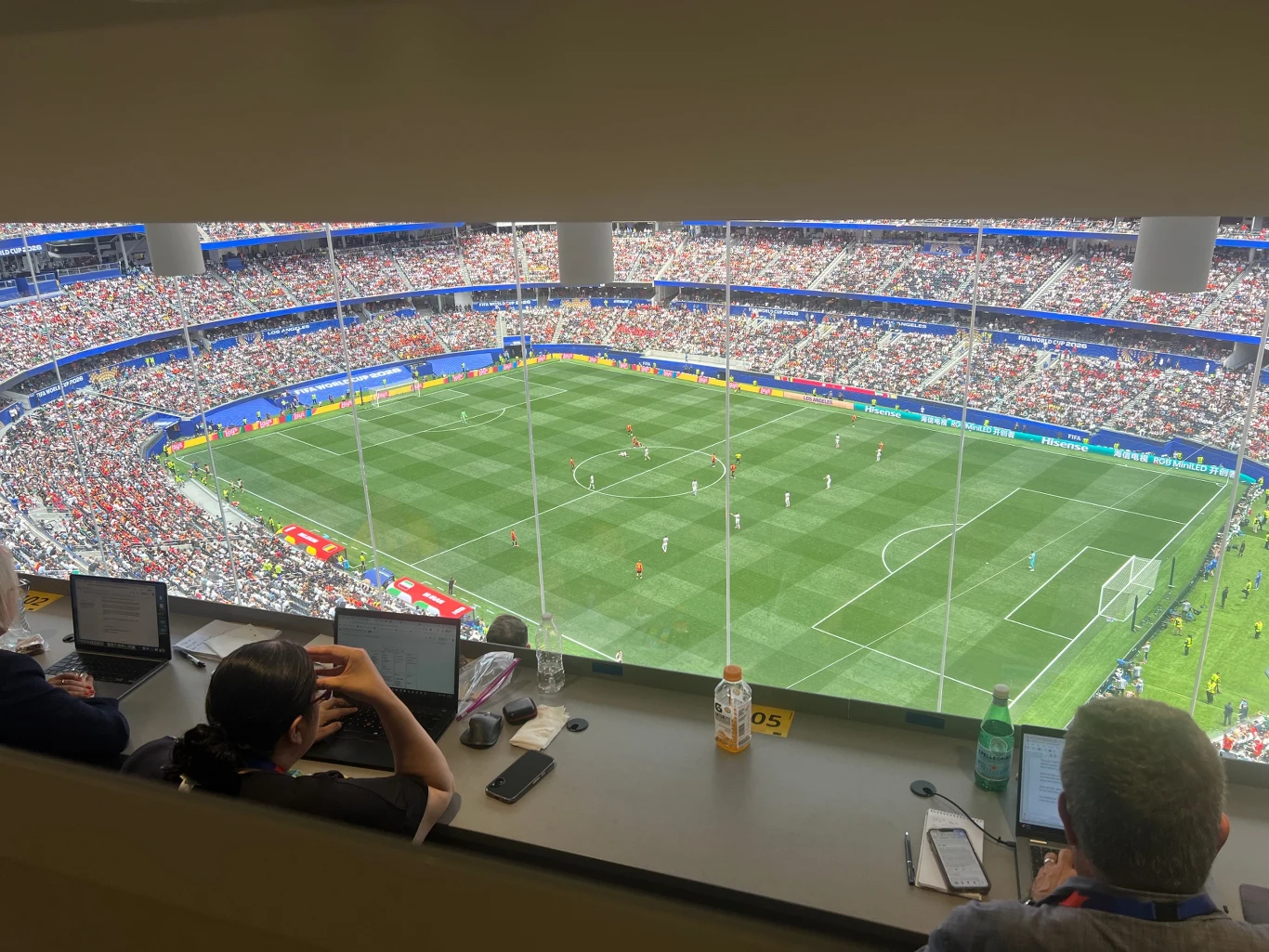 Widok na pełny stadion podczas meczu piłki nożnej, trybuny wypełnione kibicami, w loży prasowej kilku dziennikarzy pracujących przy komputerach.