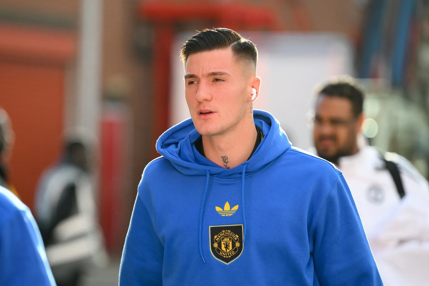 Młody mężczyzna w niebieskiej bluzie z kapturem z emblematem Manchester United oraz logo Adidas, spaceruje w słonecznym otoczeniu miejskim.