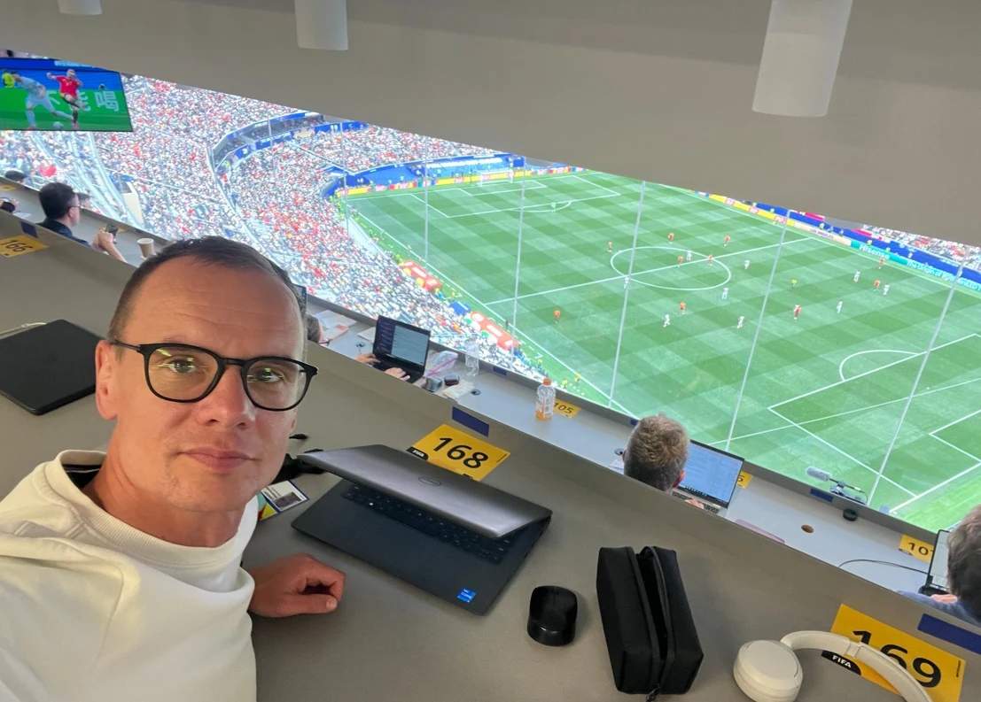Mężczyzna w okularach siedzi w loży prasowej stadionu piłkarskiego, przed nim laptop i akcesoria biurowe, w tle widok na murawę, na której rozgrywany jest mecz oraz trybuny wypełnione kibicami.
