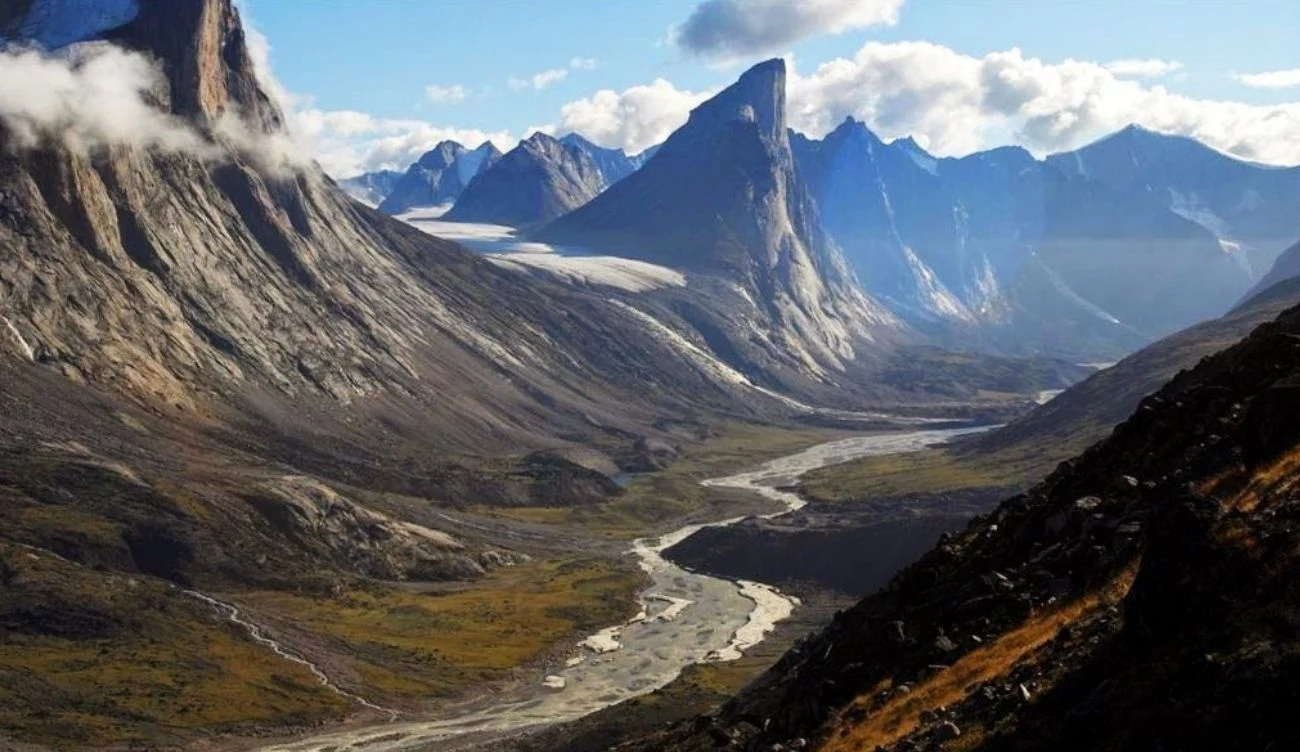 Mount Thor jest najwyższą pionową ścianą na świecie Szczyt Mount Thor z pionową ścianą, otoczony górami i doliną z wijącą się rzeką oraz pasmami zieleni.