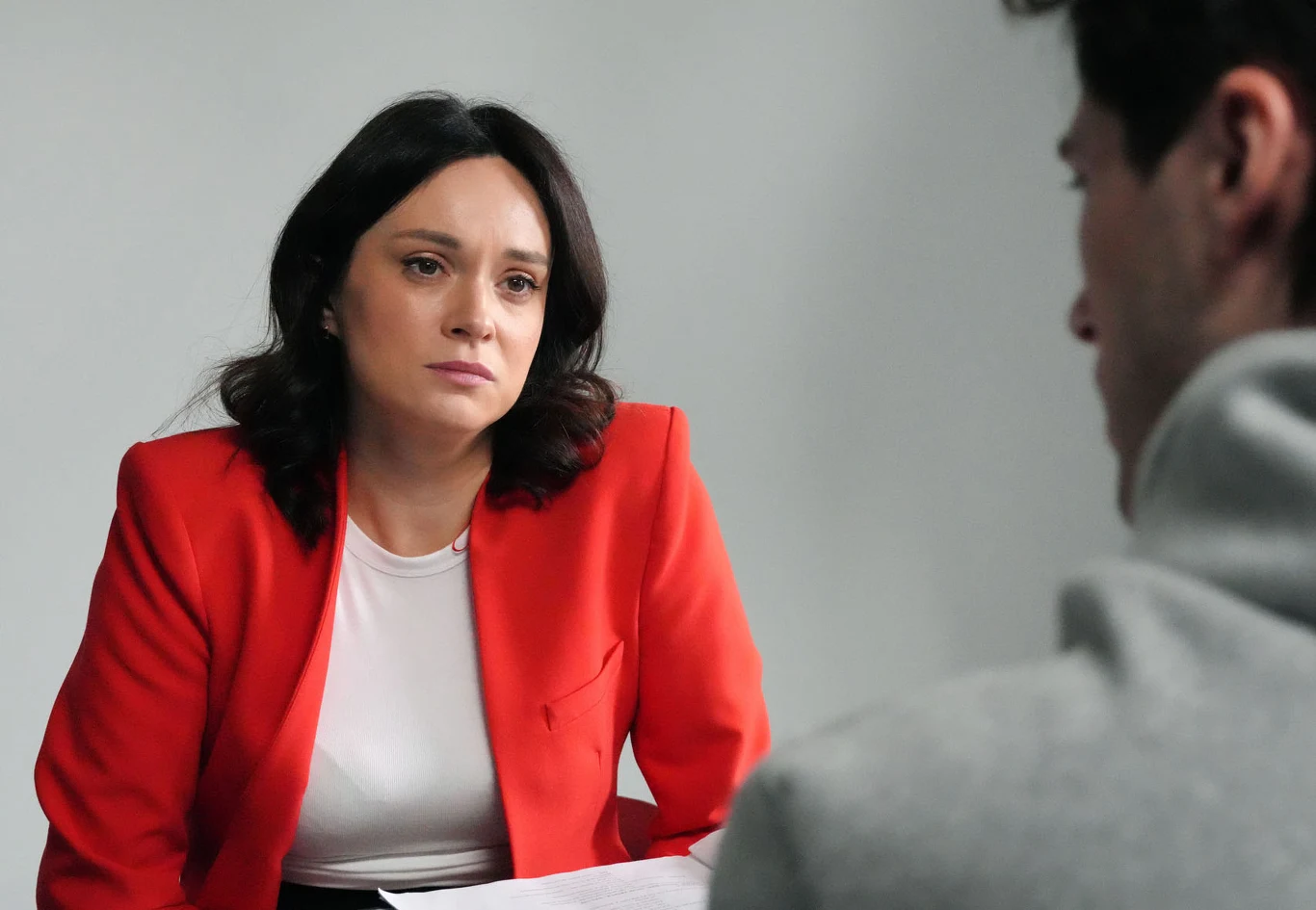 Kobieta w czerwonej marynarce siedzi przy stole naprzeciw mężczyzny, patrząc na niego z powagą.
