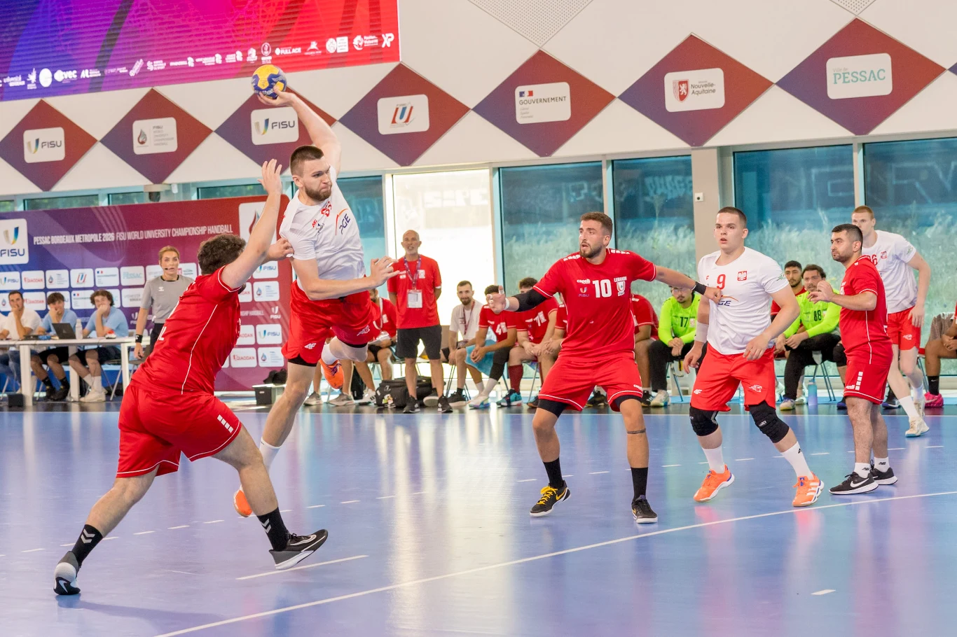 Zawodnik w białej koszulce wyskakuje do rzutu piłką podczas meczu piłki ręcznej, otoczony przez kilku obrońców w czerwonych strojach, próbujących go zablokować.