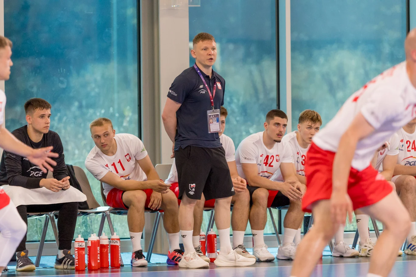 Grupa młodych szczypiornistów w biało-czerwonych strojach siedzi na ławce rezerwowych z trenerem na środku, skoncentrowanym na przebiegu meczu, podczas gdy jeden z zawodników w trakcie gry pojawia się na pierwszym planie.