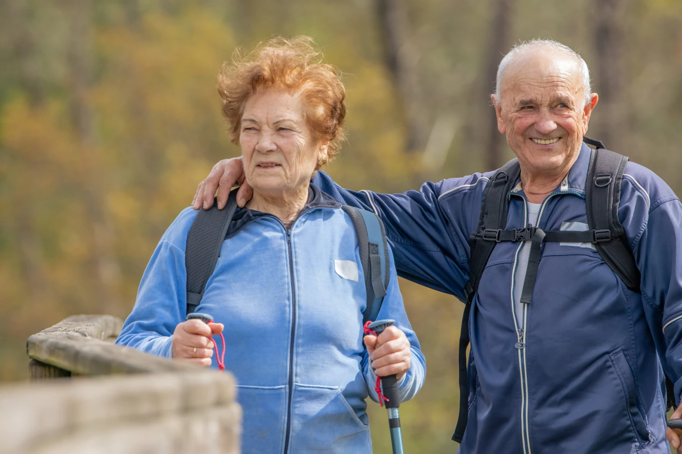 Starsze małżeństwo w sportowych ubraniach spaceruje razem na świeżym powietrzu, oboje mają plecaki i kijki trekkingowe, mężczyzna obejmuje kobietę ramieniem, oboje wyglądają na zrelaksowanych i szczęśliwych.