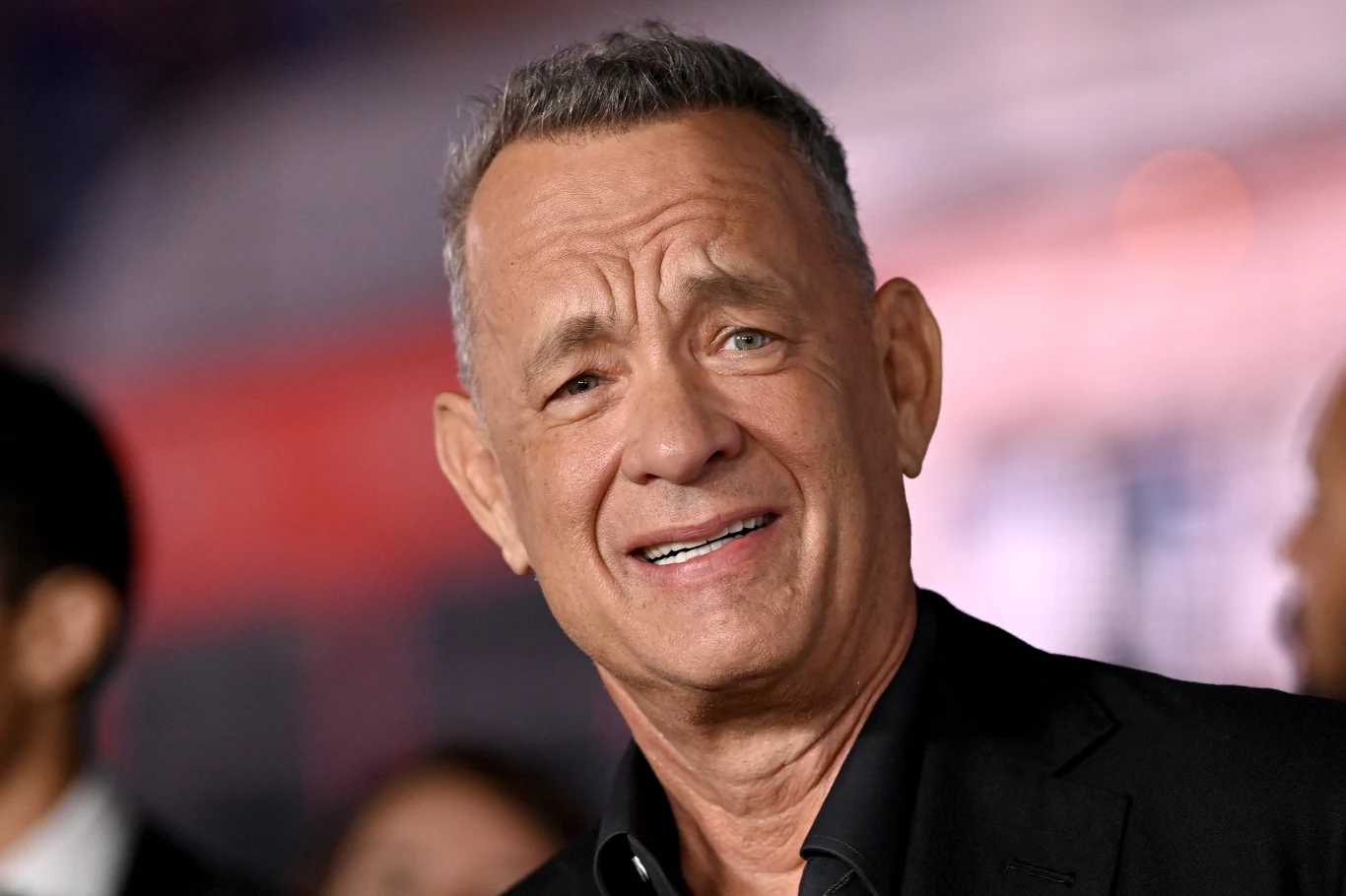 Tom Hanks Tom Hanks z siwymi włosami w czarnej marynarce, lekko uśmiechnięty, na rozmytym, kolorowym tle.