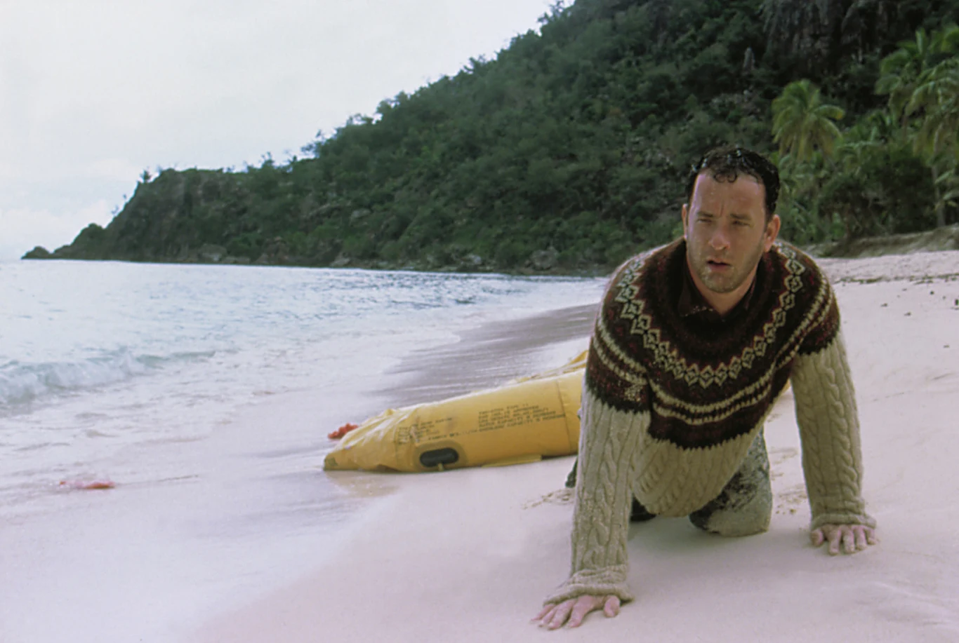 Tom Hanks w filmie "Cast Away: Poza światem" Zasapany mężczyzna w swetrze klęczy na piaszczystej plaży obok fragmentu żółtej tratwy, a w tle rozciąga się gęsto zalesiony brzeg tropikalnej wyspy.