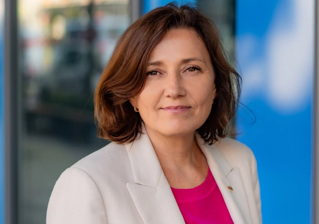 Kobieta w jasnej marynarce i różowej bluzce patrząca w stronę obiektywu, o delikatnym uśmiechu, na tle niebiesko-szklanej powierzchni.