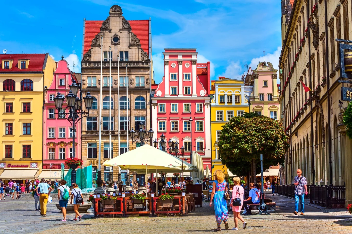 Kolorowe kamienice z efektowną fasadą, ogródek kawiarniany z dużym parasolem w centrum placu oraz spacerujący ludzie na rynku miejskim w słoneczny dzień.