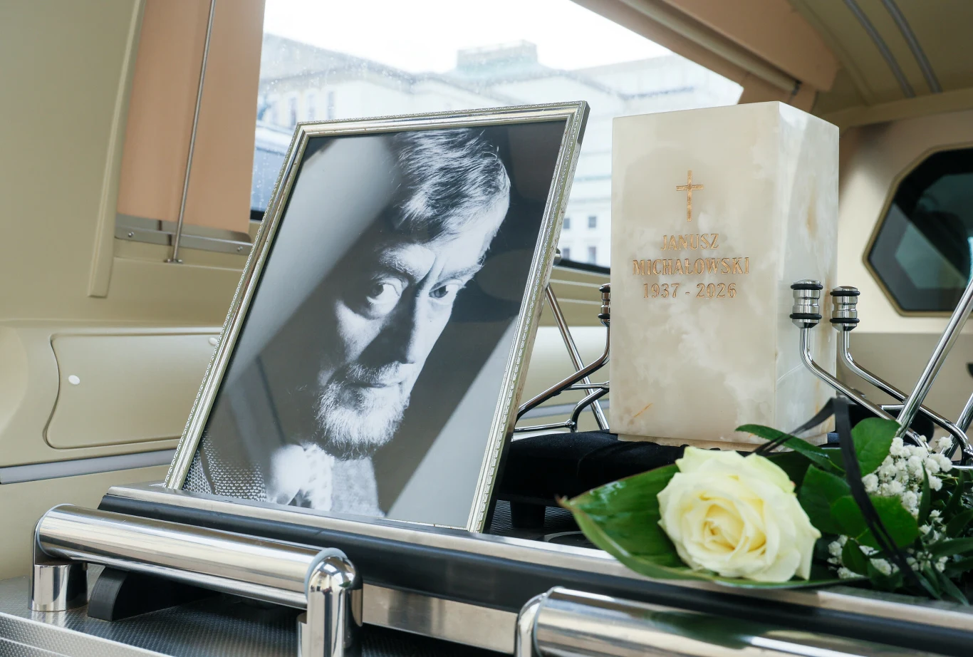 Pogrzeb Janusza Michałowskiego Urna z prochami Janusza Michałowskiego z białą różą oraz zdjęcie aktora w srebrnej ramie podczas uroczystości pogrzebowych.