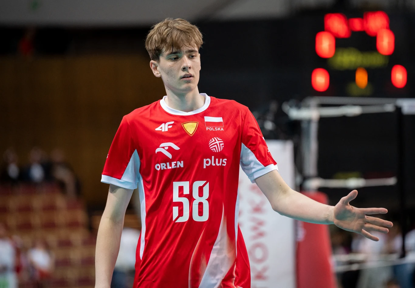 Bartosz Zych Młody siatkarz w czerwonym stroju reprezentacji Polski z numerem 58 na piersi podczas meczu, na tle rozmytej hali sportowej i widocznej tablicy wyników.
