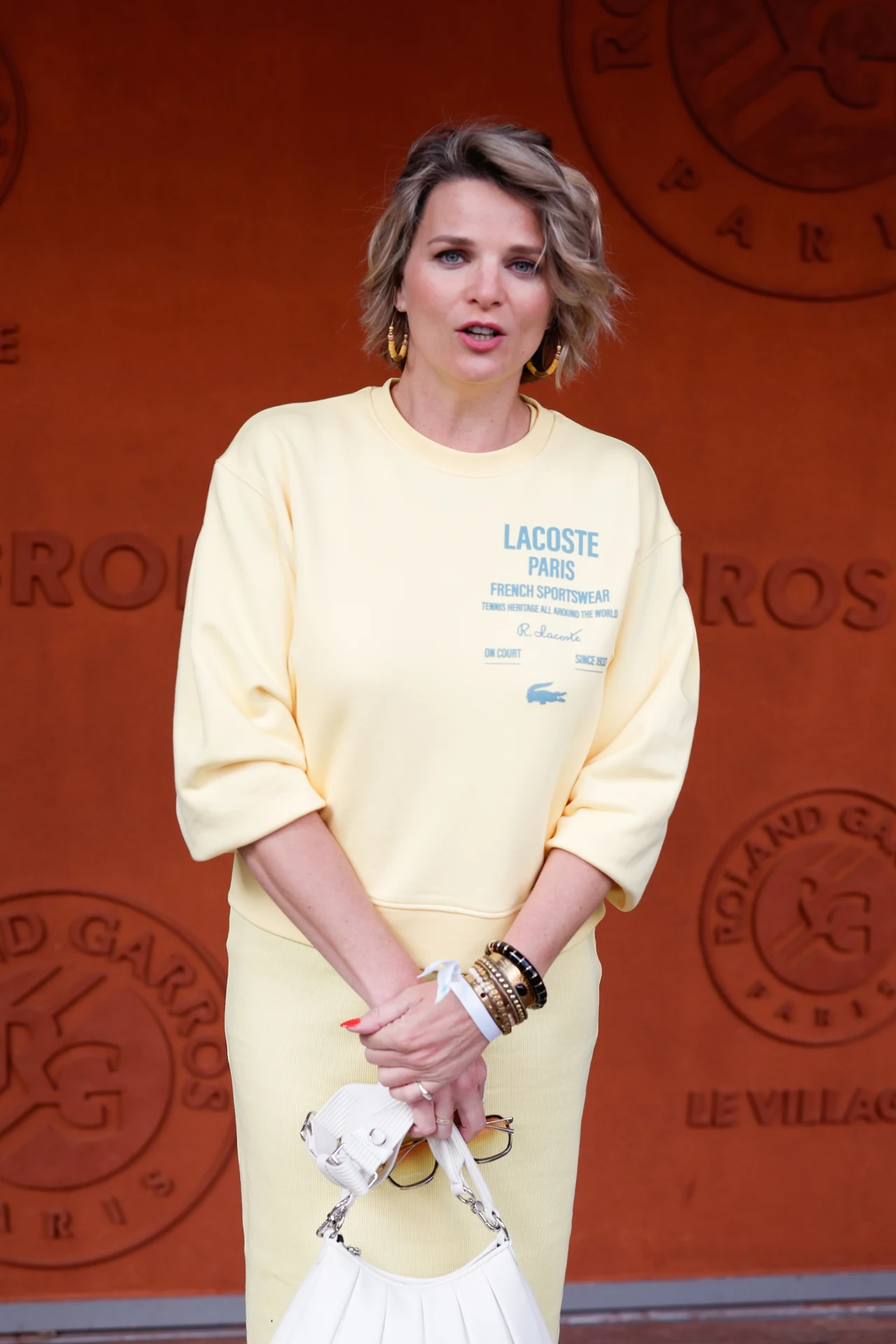 France Pierron Kobieta w jasnożółtej sukience z nadrukiem Lacoste trzyma białą torebkę, stoi na tle czerwonej ściany z logo Roland Garros.