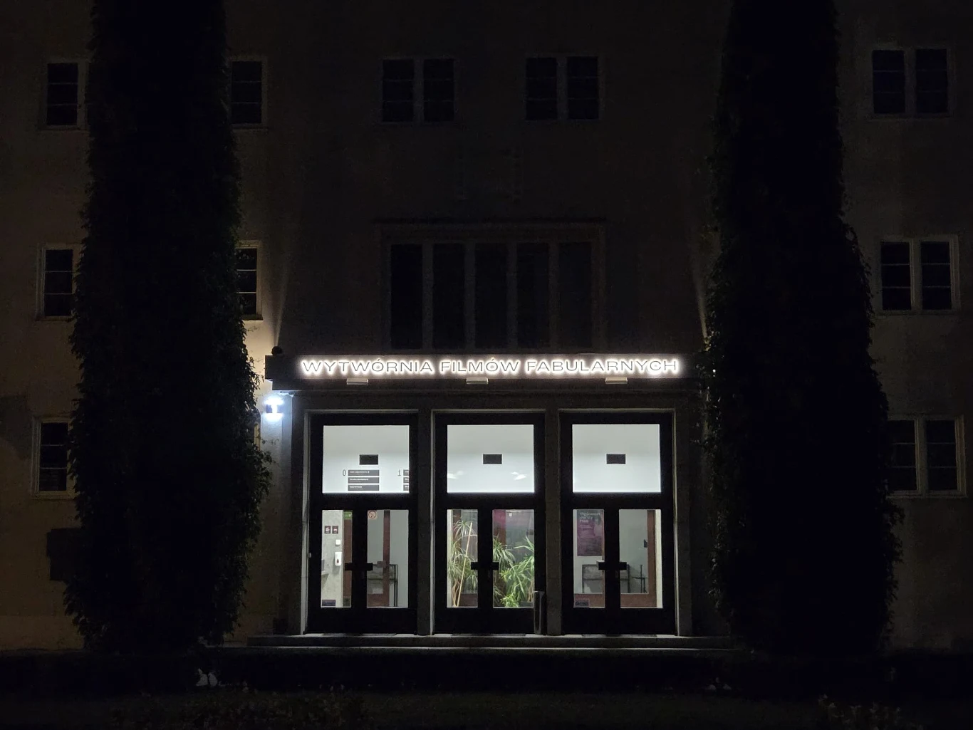 Oświetlony neon z napisem „Wytwórnia Filmów Fabularnych” nad wejściem do budynku, otoczonym przez dwie wysokie drzewa po bokach.