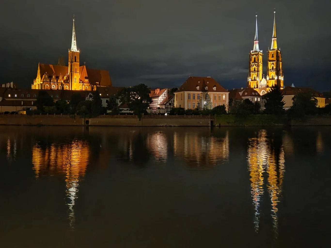 Oświetlona nocą katedra oraz kościół odbijające się w spokojnej tafli rzeki, otoczone niską zabudową miejską.