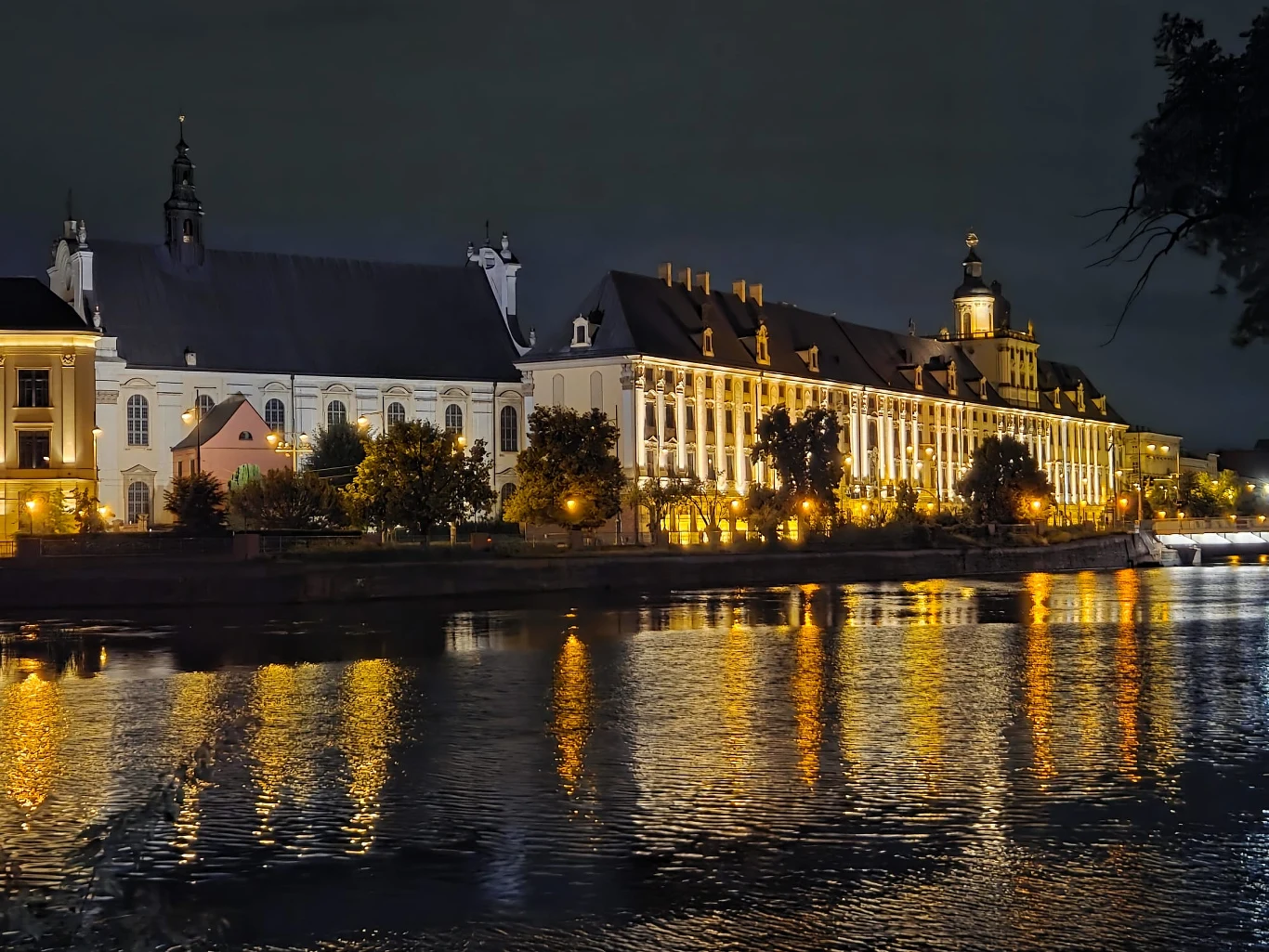 Oświetlone budynki w stylu barokowym odbijające się w spokojnej tafli rzeki nocą.