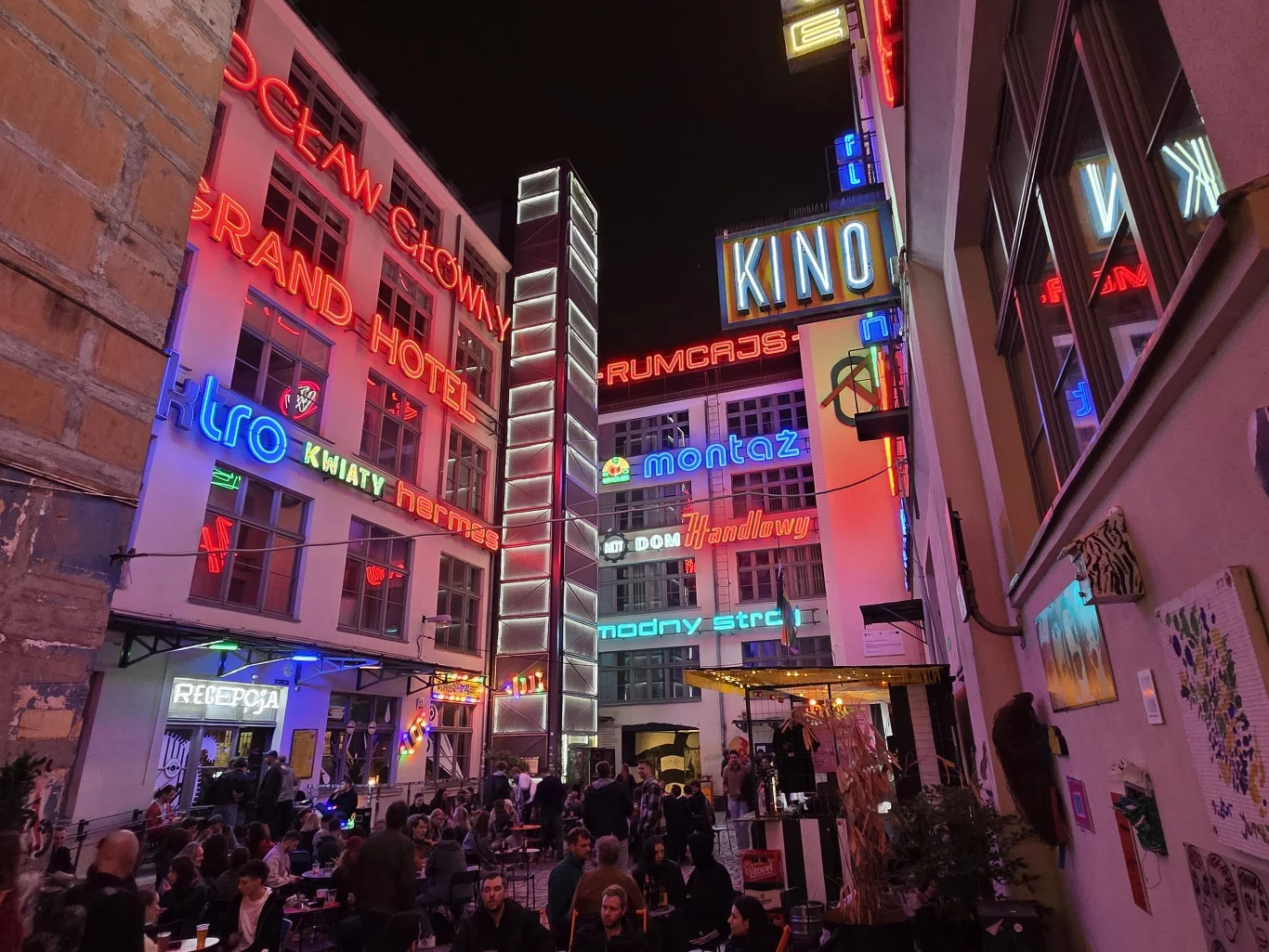 Kolorowe neony z napisami w różnych stylach rozświetlają nocą miejskie podwórko, wokół którego gromadzą się ludzie siedzący w kawiarnianych ogródkach.