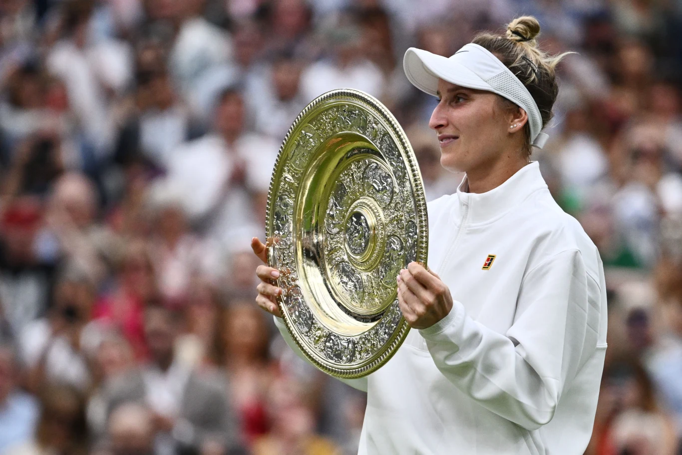 Sportowiec ubrany w białą bluzę i opaskę na głowie trzyma dużą, ozdobną, złotą tacę z wygrawerowanymi wzorami na tle rozmytego tłumu na trybunach.