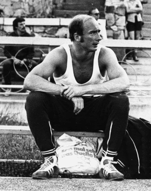Mężczyzna w sportowym stroju siedzi na ławce z wyprostowanymi łokciami opartymi na kolanach, przy nogach leży torba sportowa, w tle widoczne trybuny i kilku ludzi siedzących na ławkach.