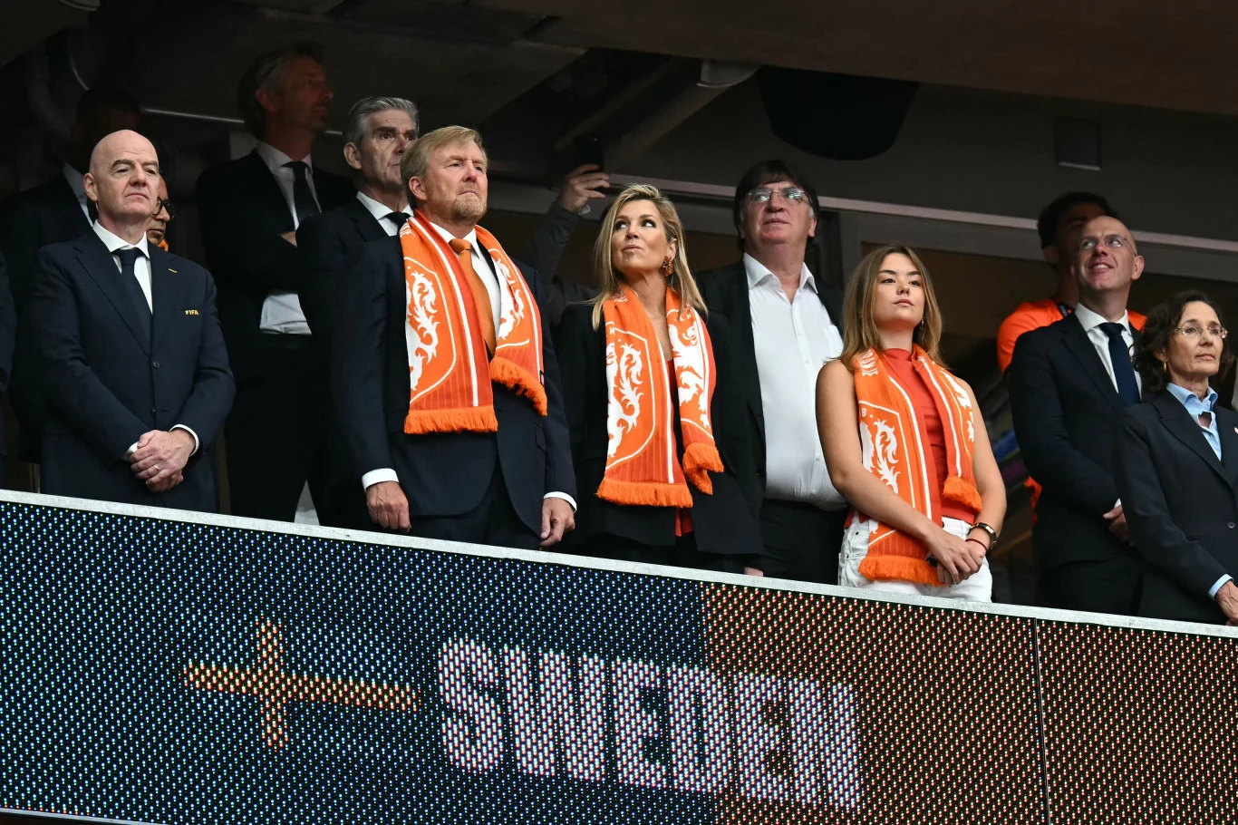 Grupa elegancko ubranych osób w pomarańczowych szalikach z godłem Holandii stoi na trybunach stadionu podczas meczu piłkarskiego ze Szwecją.