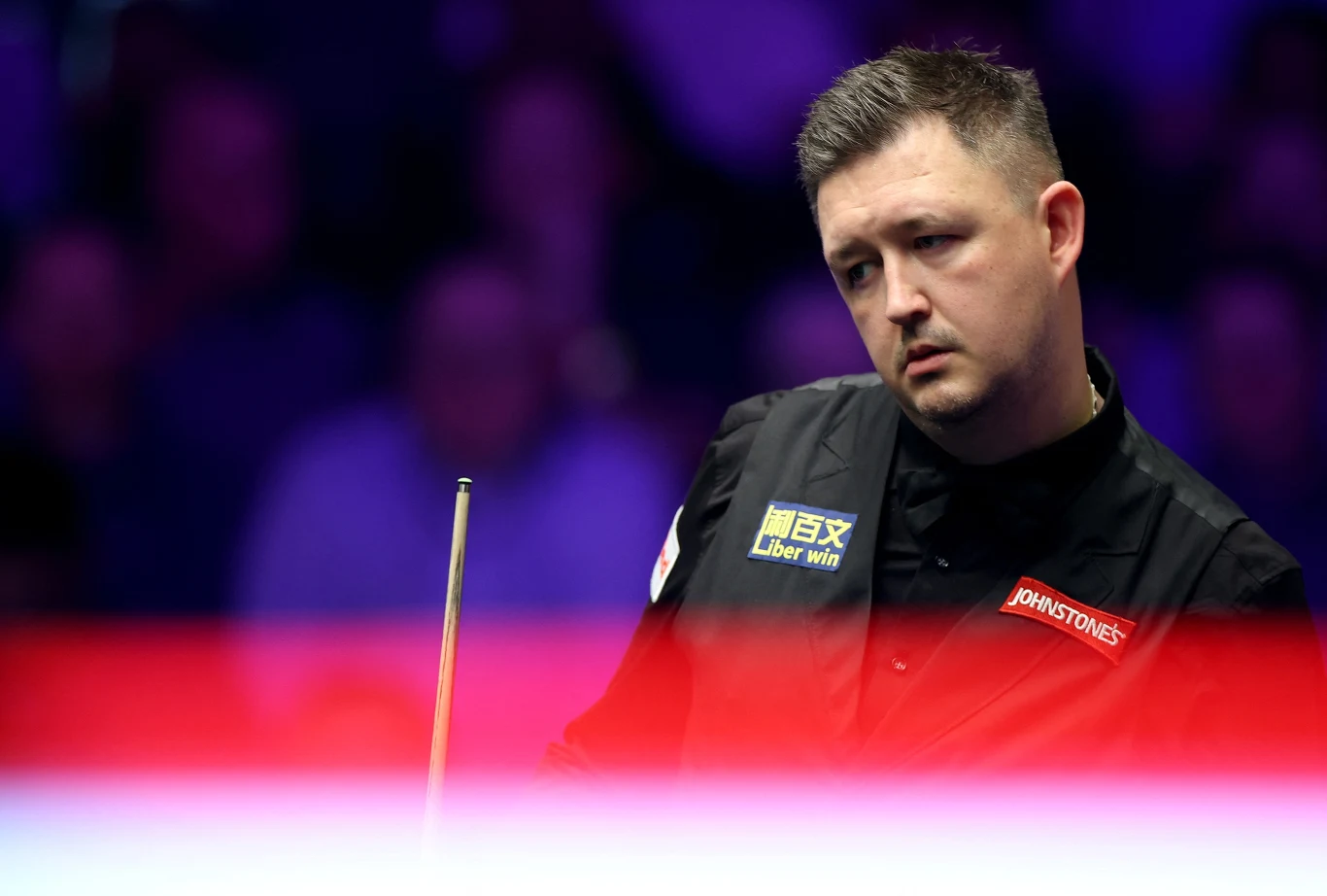 Zawodowy snookerzysta w ciemnej kamizelce z kijem bilardowym w ręku, skupiony nad rozgrywką przy oświetlonym stole podczas turnieju.