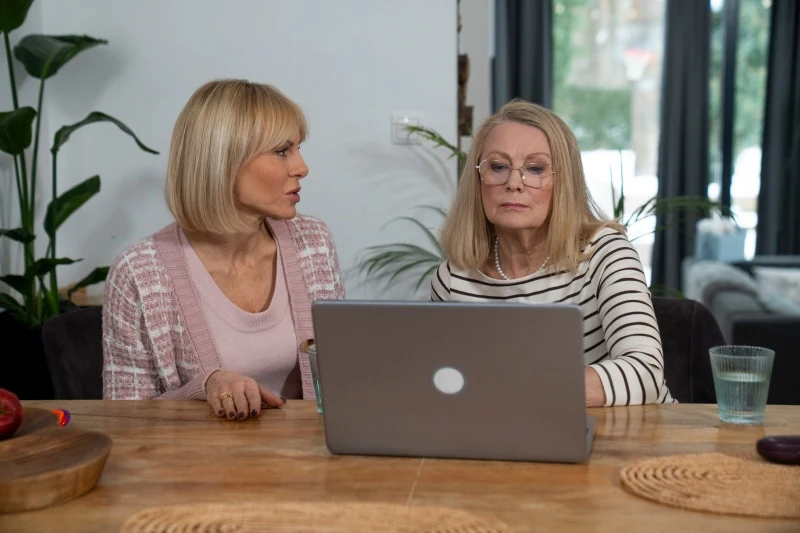 Dwie kobiety siedzą przy drewnianym stole, skupione na ekranie otwartego laptopa; obok stoi szklanka z wodą.