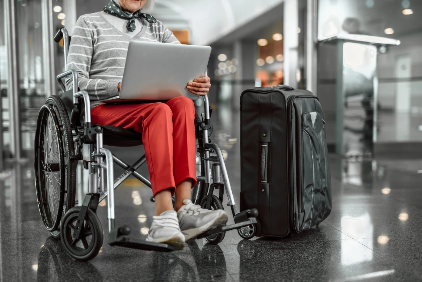 Osoba na wózku inwalidzkim w jasnym swetrze i czerwonych spodniach korzysta z laptopa, obok stoi duża czarna walizka podróżna.