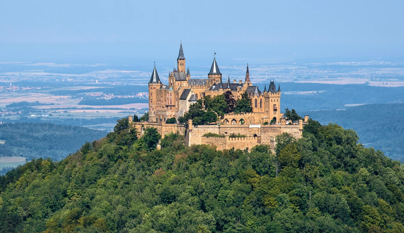 Gdzie jest zamek Hohenzollern? Zamek Hohenzollern z wieloma wieżami na zalesionym wzgórzu, otoczony zielenią, z widokiem na płaską okolicę.
