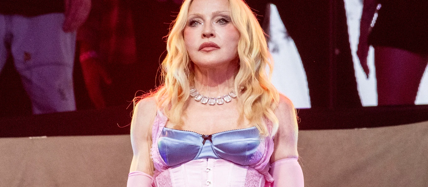 Madonna Madonna w jasnoróżowym gorsecie i niebieskim biustonoszu, stoi na scenie w ciemnym otoczeniu z innymi postaciami.