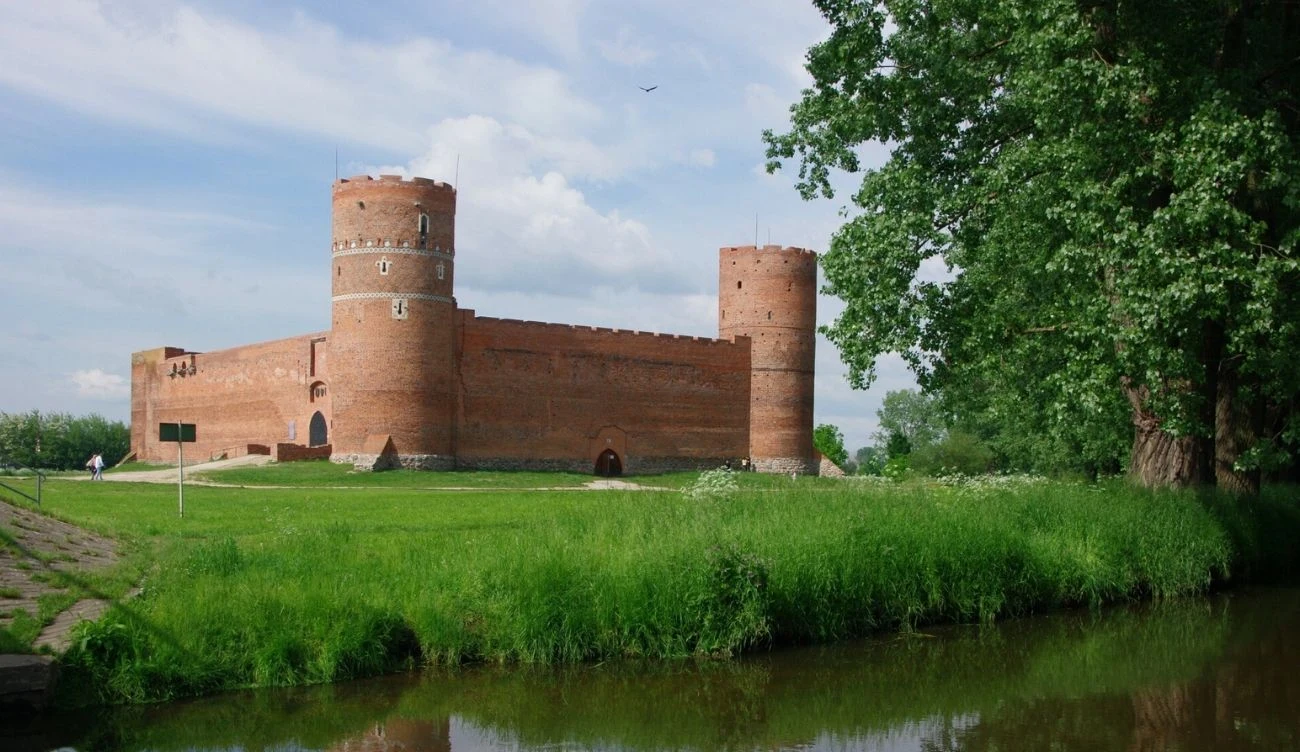 Zamek Książąt Mazowieckich przez wieki pozostawał niezdobyty Ceglany Zamek Książąt Mazowieckich w Ciechanowie z basztami cylindrycznymi, w otoczeniu zieleni i z widokiem na rzekę.