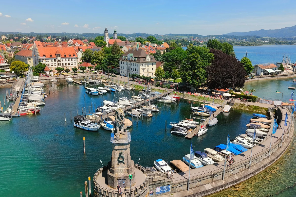 Port z przystanią jachtową otoczony mieszkalnymi i zabytkowymi budynkami, w centralnym miejscu masywna kamienna statua lwa, wokół zielone drzewa oraz turkusowa woda jeziora.