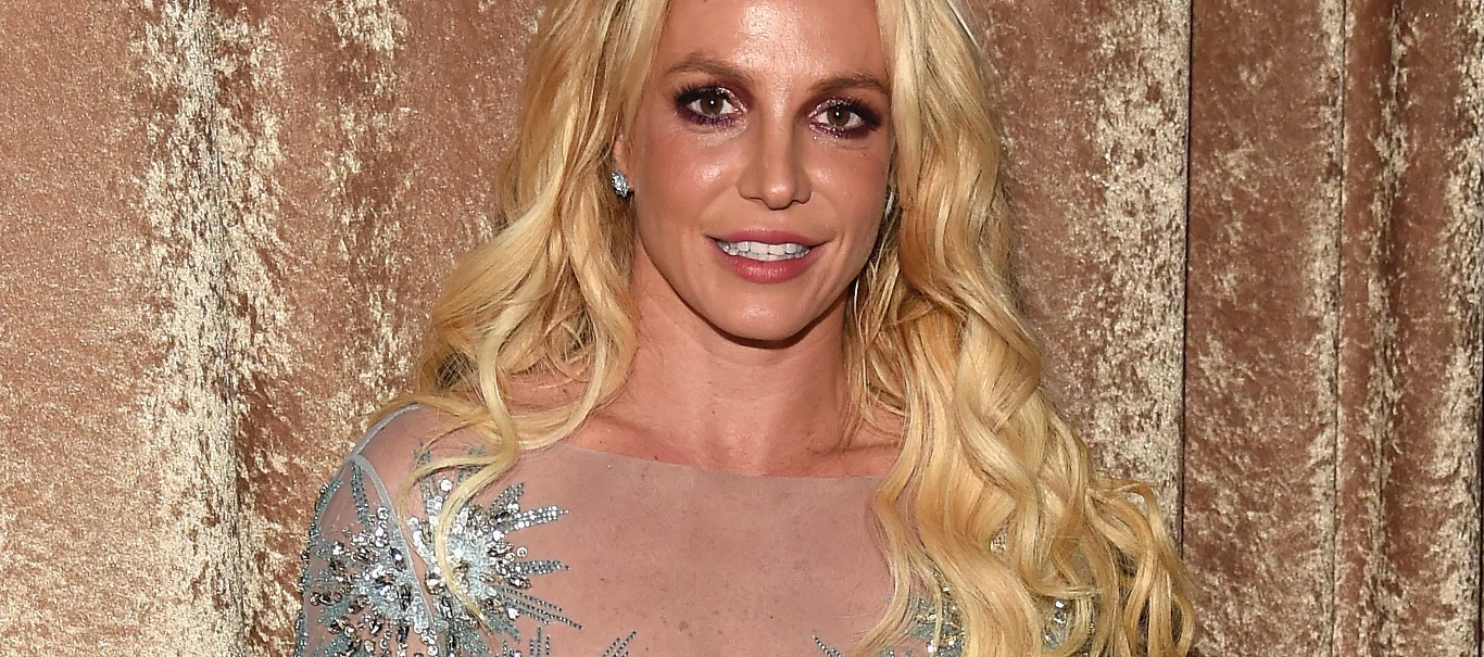 Britney Spears Britney Spears w długich blond włosach, w niebieskiej cekinowej sukni na tle złotobrązowej zasłony.