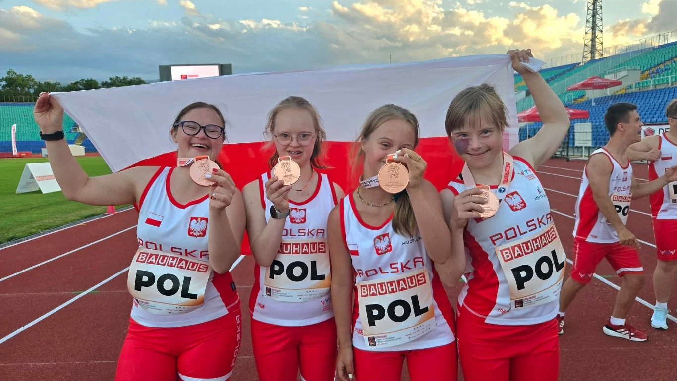 Cztery młode lekkoatletki w biało-czerwonych strojach z napisem POL trzymają medale i polską flagę, świętując wspólny sukces na stadionie lekkoatletycznym.