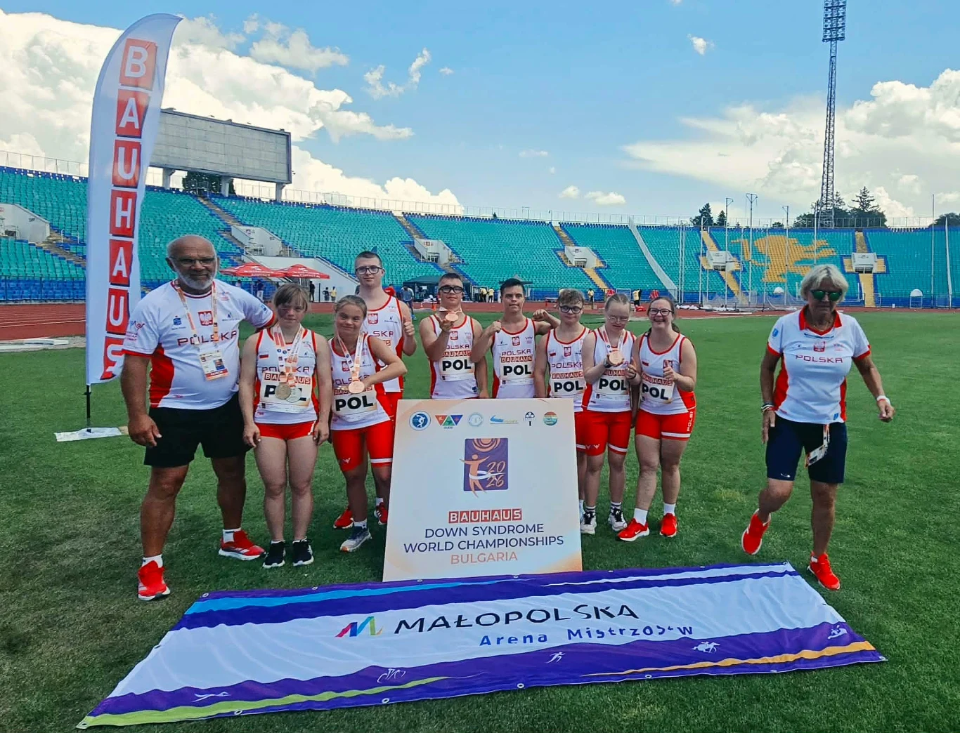 Uśmiechnięta drużyna lekkoatletyczna w biało-czerwonych strojach z napisem POL pozująca na stadionie przy tablicy informującej o mistrzostwach świata osób z zespołem Downa w Bułgarii, obok baneru Małopolska Arena Mistrzów.