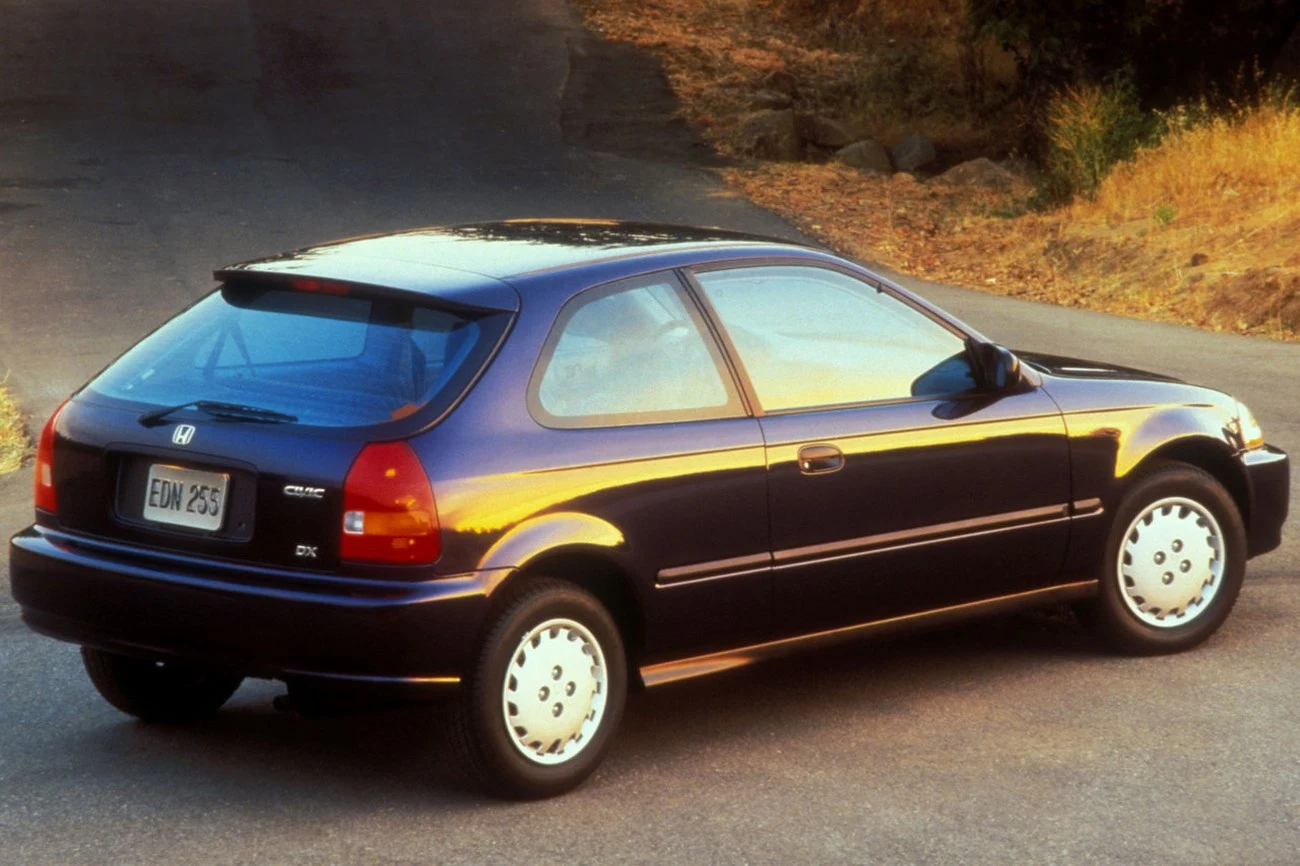 Granatowy hatchback Honda Civic z lat 90., stojący na asfaltowej drodze, oświetlony promieniami zachodzącego słońca, wyróżniający się charakterystycznym kształtem tylnej części nadwozia i dużymi tylnymi światłami.