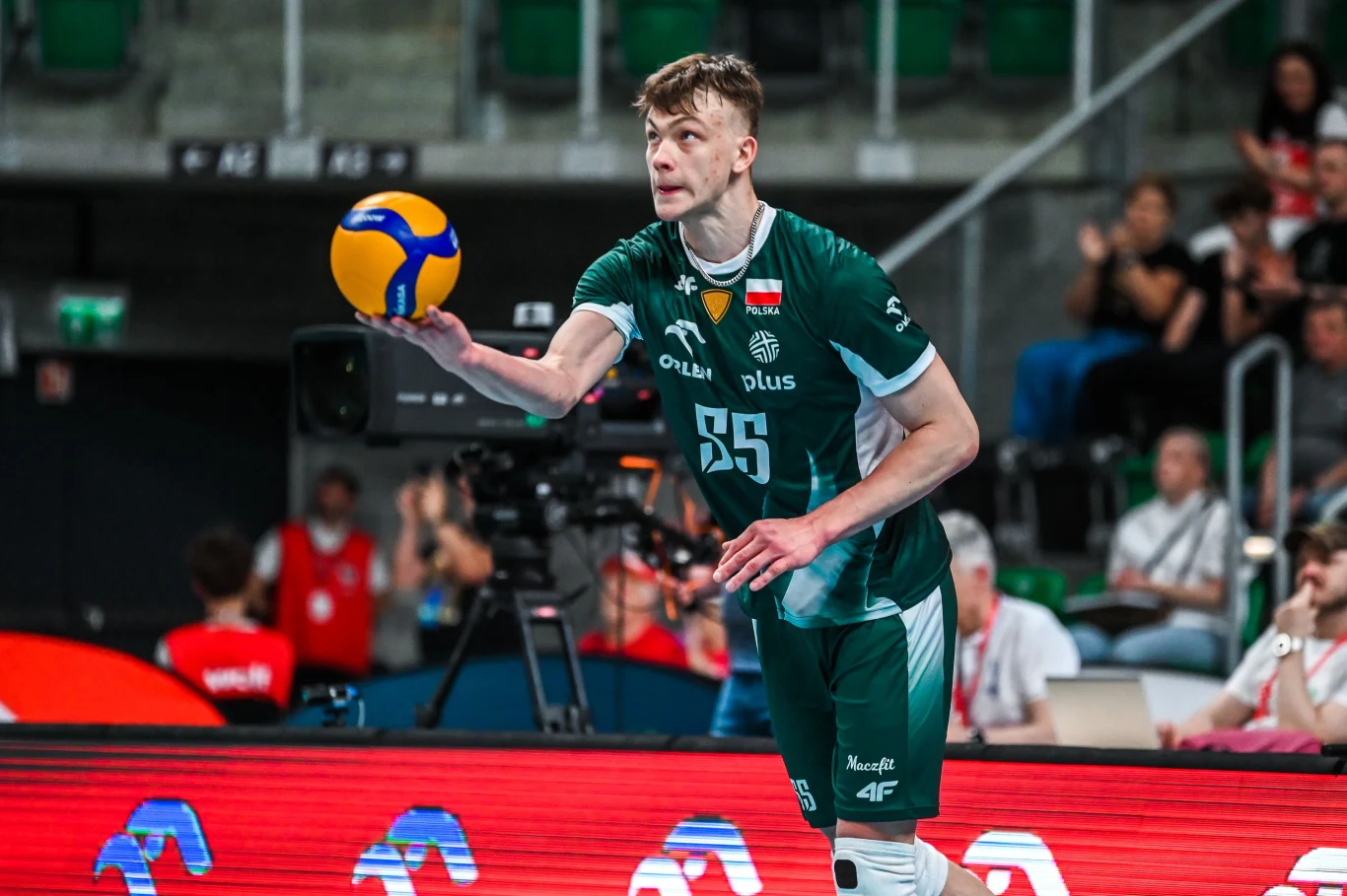 Jakub Kiedos Siatkarz w zielonym stroju przygotowujący się do wykonania zagrywki podczas meczu, z piłką uniesioną w lewej ręce i skupionym wyrazem twarzy.