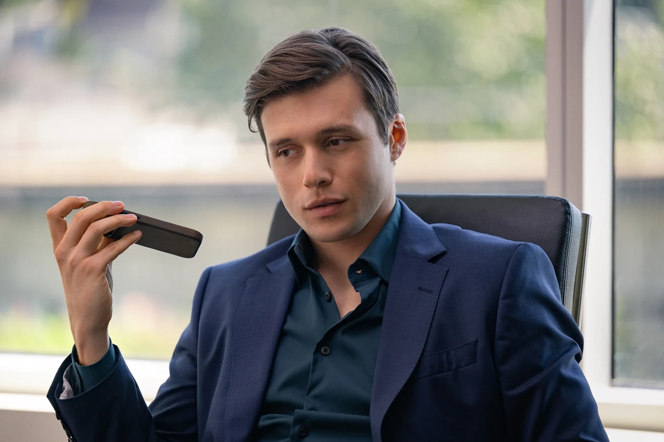 Młody mężczyzna w granatowym garniturze siedzi na krześle biurowym, trzyma telefon przy ustach i spogląda w dal z poważnym wyrazem twarzy.