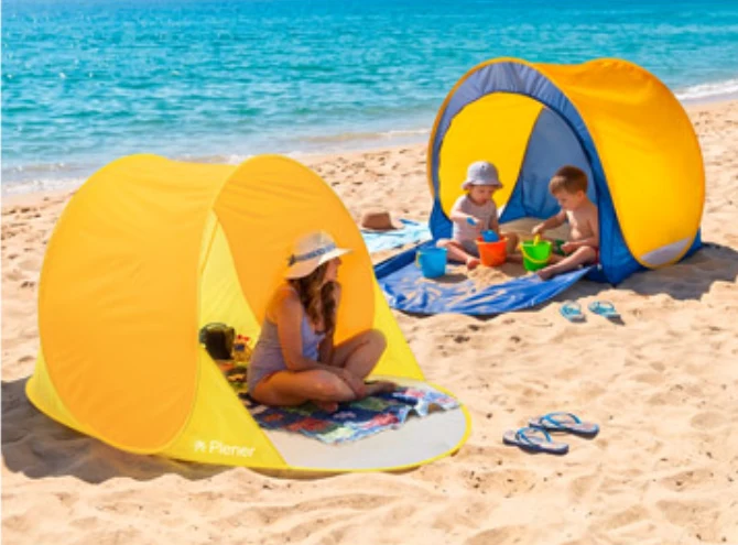 Dwie osoby dorosłe oraz dwoje dzieci siedzą w plażowych namiotach na żółtym piasku blisko brzegu morza; dzieci bawią się wiaderkami i łopatką, a wszyscy mają na głowach letnie kapelusze chroniące przed słońcem.