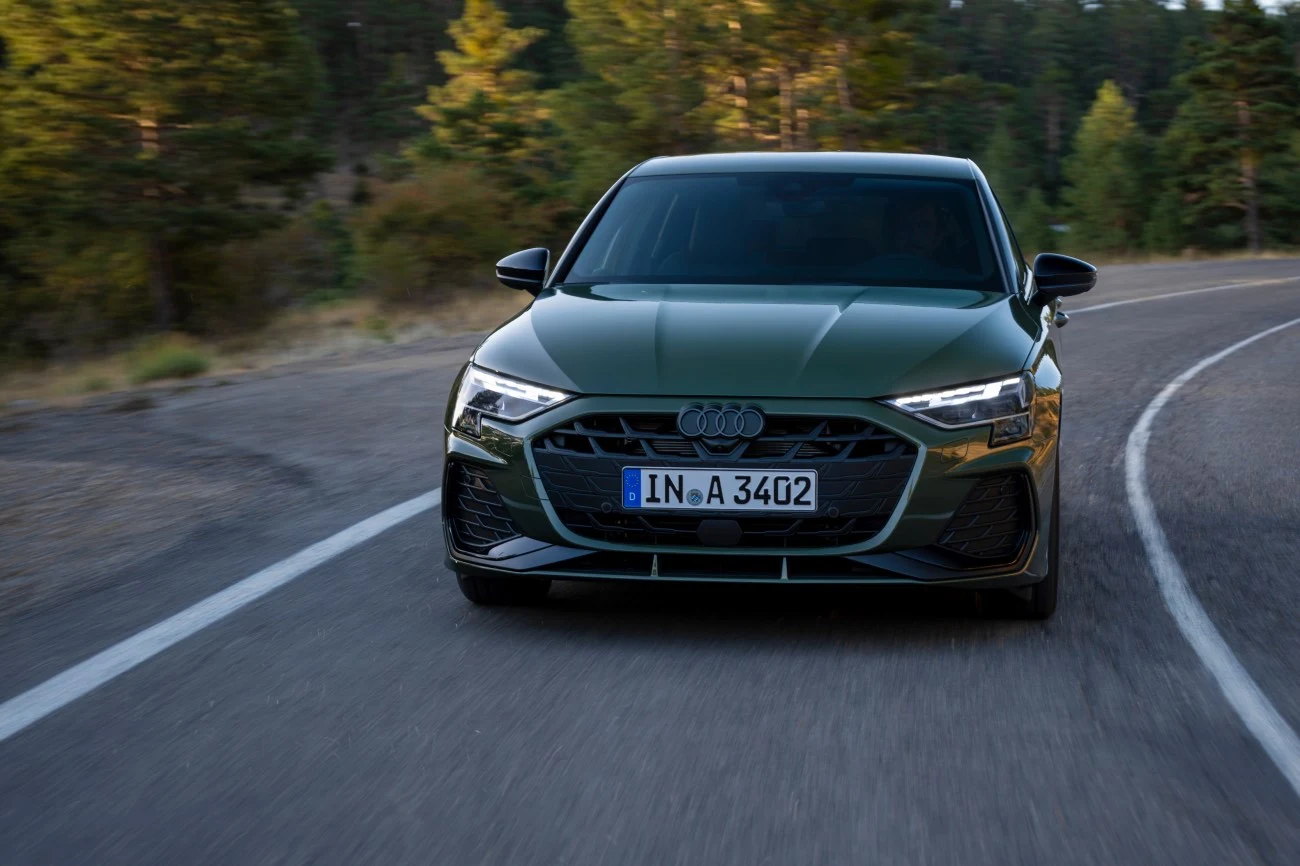 ciemnozielone Audi A3 z charakterystycznym grillem jedzie pustą, asfaltową drogą otoczoną drzewami