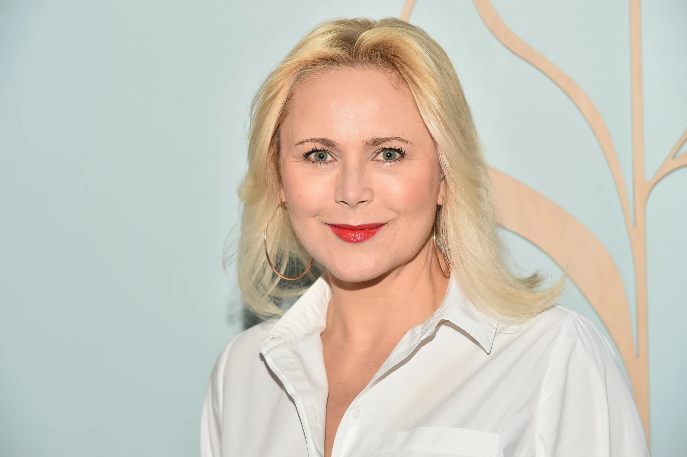 Uśmiechnięta kobieta o blond włosach ubrana w białą koszulę na tle jasnej, minimalistycznej ściany z dekoracyjnym motywem.