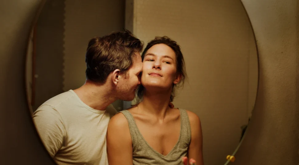 Helga Guren i Oddgeir Thune w scenie z filmu "Być kochaną" Para obejmuje się blisko przed lustrem, mężczyzna całuje policzek kobiety, a ona delikatnie się uśmiecha.