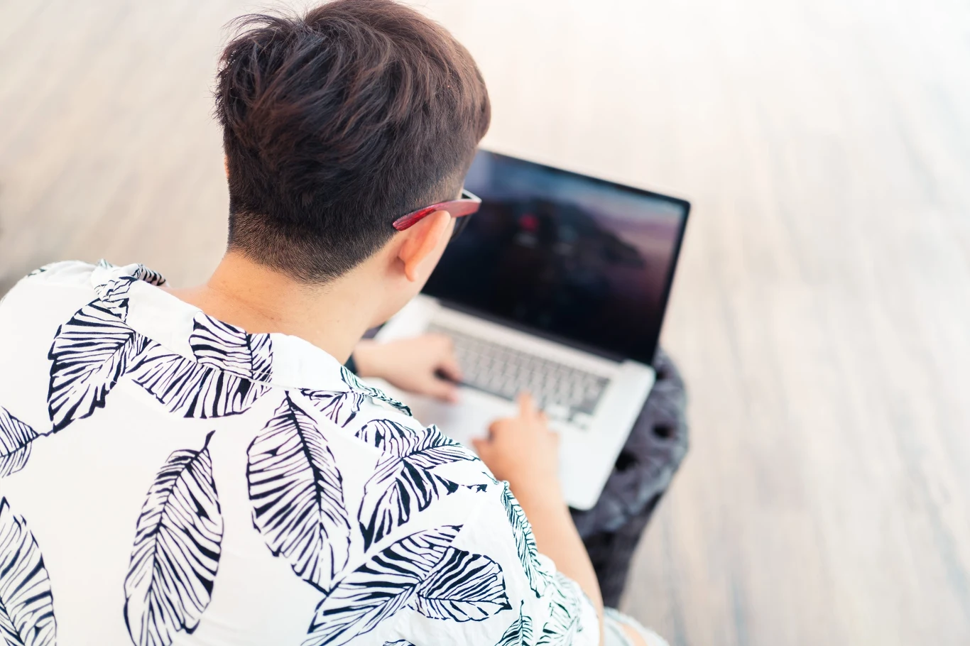 Mężczyzna w okularach i koszuli w liście pracuje na laptopie, siedząc na krześle na jasnej powierzchni.