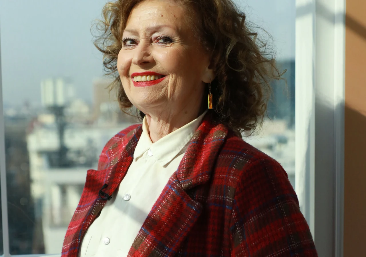 Ewa Florczak Ewa Florczak w białej koszuli i czerwonej marynarce w kratę na tle okna z widokiem na miasto.