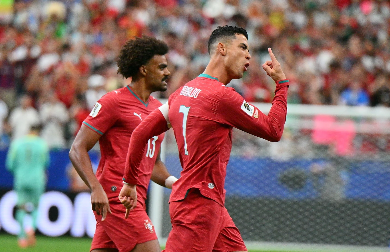 Cristiano Ronaldo na liście strzelców. Czekał na ten moment cierpliwie Piłkarz w czerwonym stroju z numerem 7 na plecach wykonuje gest palcem przy ustach w geście triumfu, a obok niego drugi zawodnik w czerwonym stroju idzie w jego kierunku; w tle widoczne rozmyte sylwetki kibiców na trybunach.