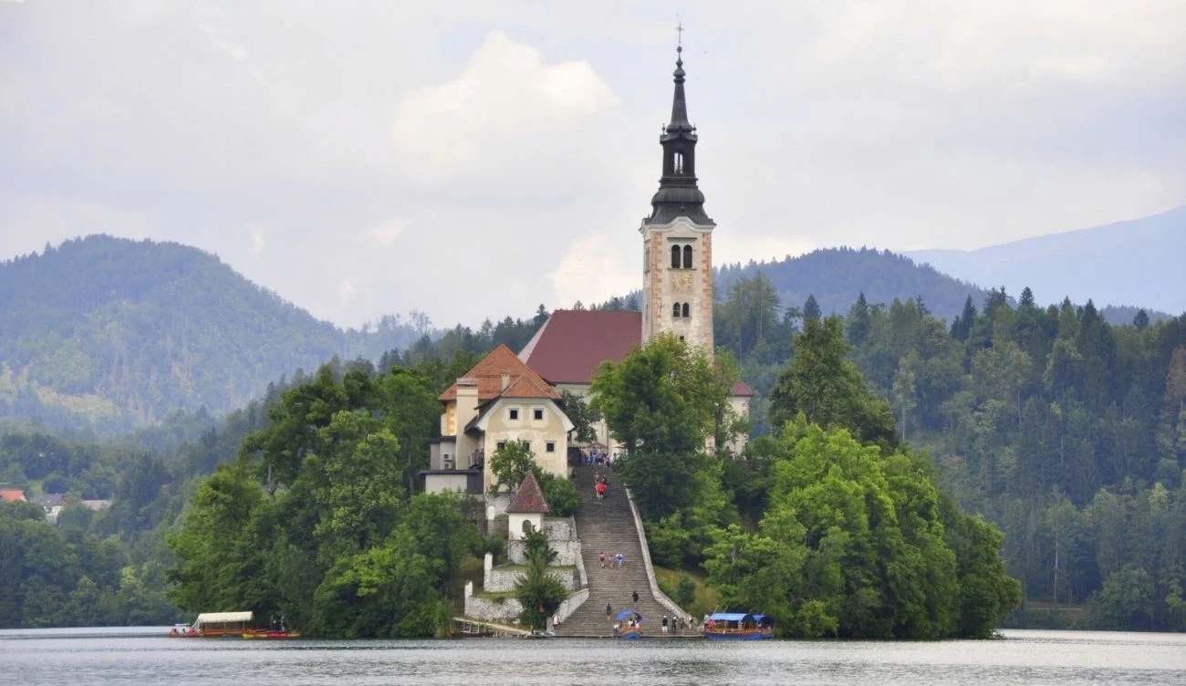 Co zobaczyć w Triglav National Park? Kościół z wysoką wieżą na zalesionej wyspie, otoczony wodą, ze schodami prowadzącymi do budynków sakralnych.