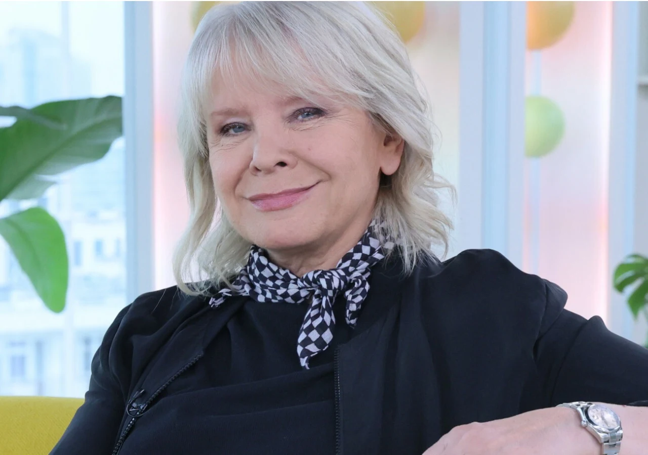 Małgorzata Domagalik Małgorzata Domagalik w czarnej marynarce i apaszce w kratkę na tle jasnego wnętrza z roślinami.