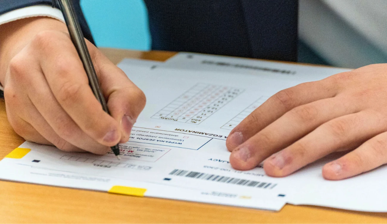 Matura 2026. Kiedy można się odwołać od wyniku egzaminu? Ręce wypełniające arkusz egzaminacyjny na stole, zbliżenie na dłoń trzymającą długopis i pola odpowiedzi.