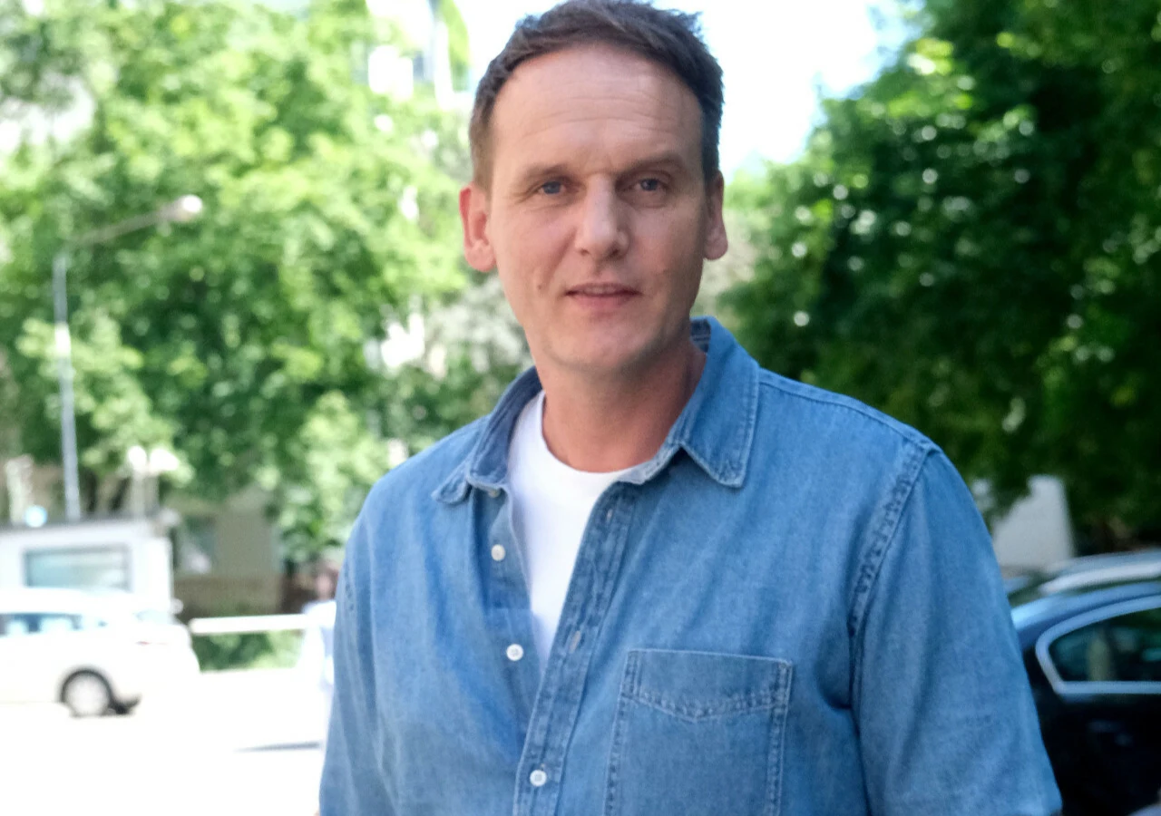 Pascal Brodnicki Pascal Brodnicki w koszuli jeansowej i białym t-shircie na tle zieleni i zaparkowanych samochodów.