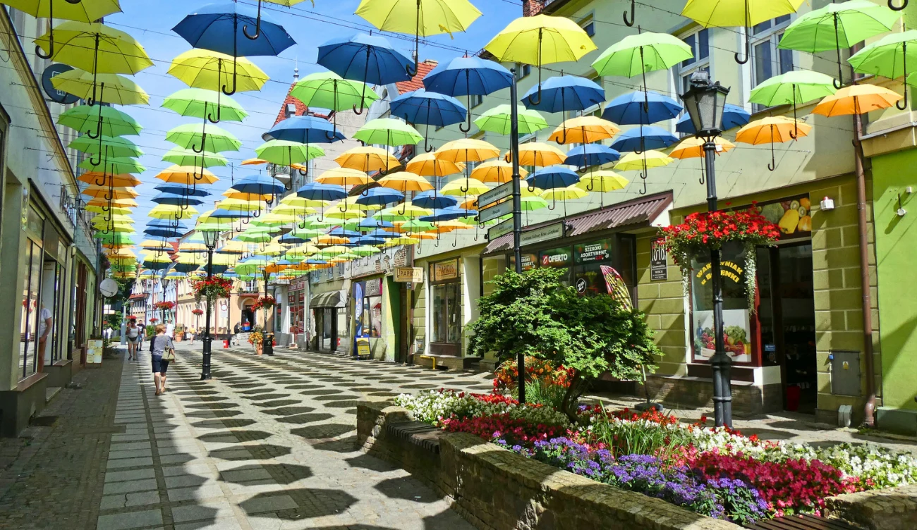 Kolorowe parasole zawieszone nad deptakiem tworzące efektowny dach, otoczone zadbanymi klombami kwiatów oraz witrynami sklepów po obu stronach ulicy.