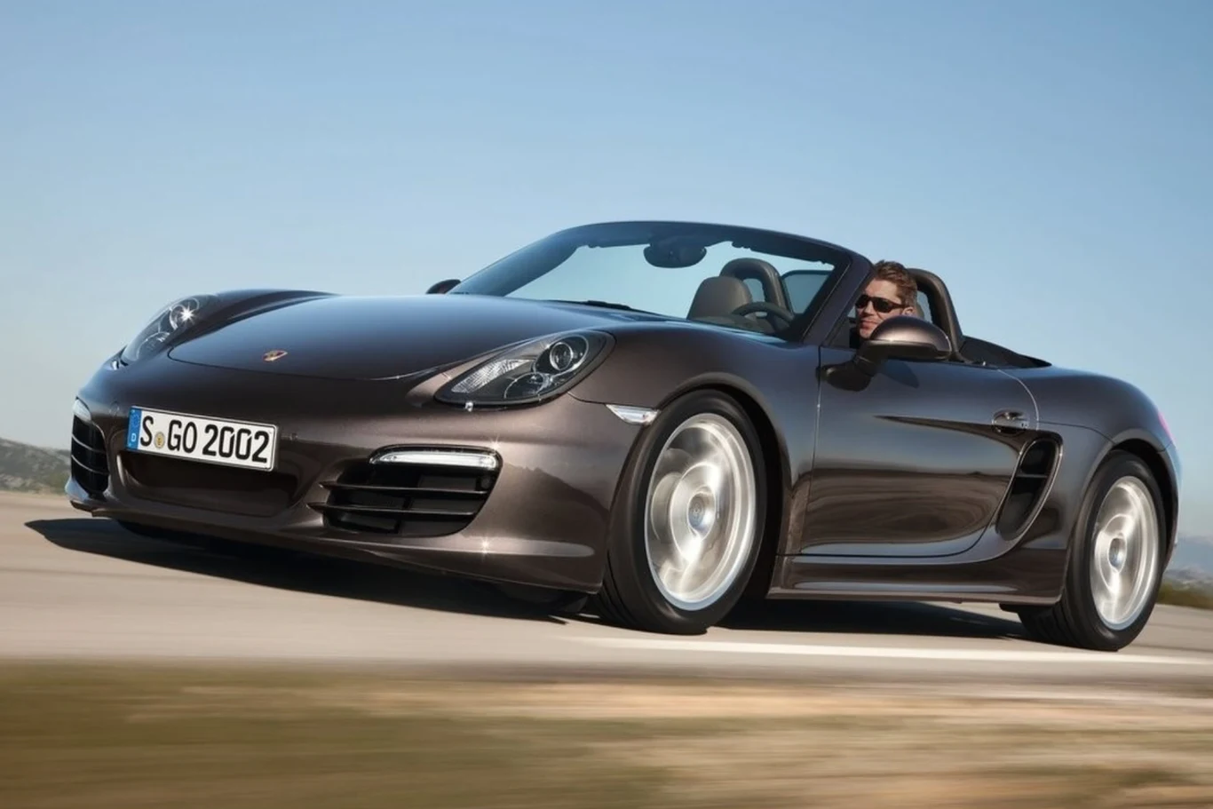 Kabriolet Porsche Boxster w ciemnym kolorze poruszający się z prędkością po asfaltowej drodze, kierowca w okularach słonecznych siedzi za kierownicą przy opuszczonym dachu.